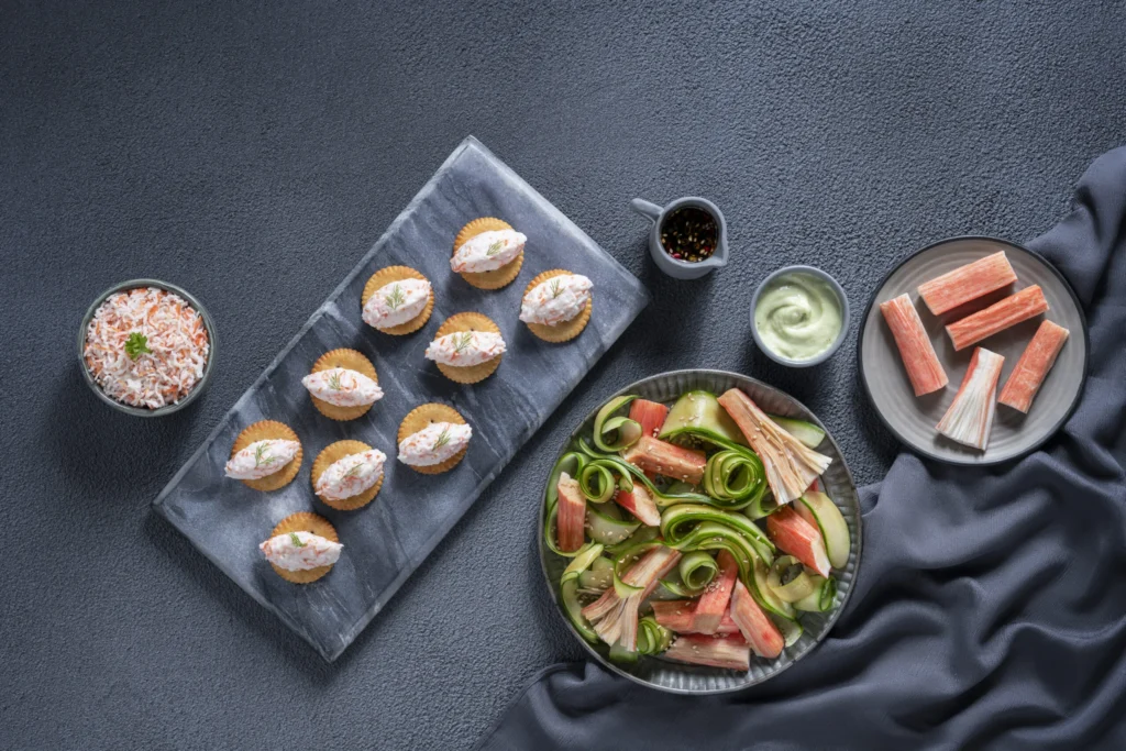 Assorted crab stick appetizers and salad arranged on a dark textured surface.