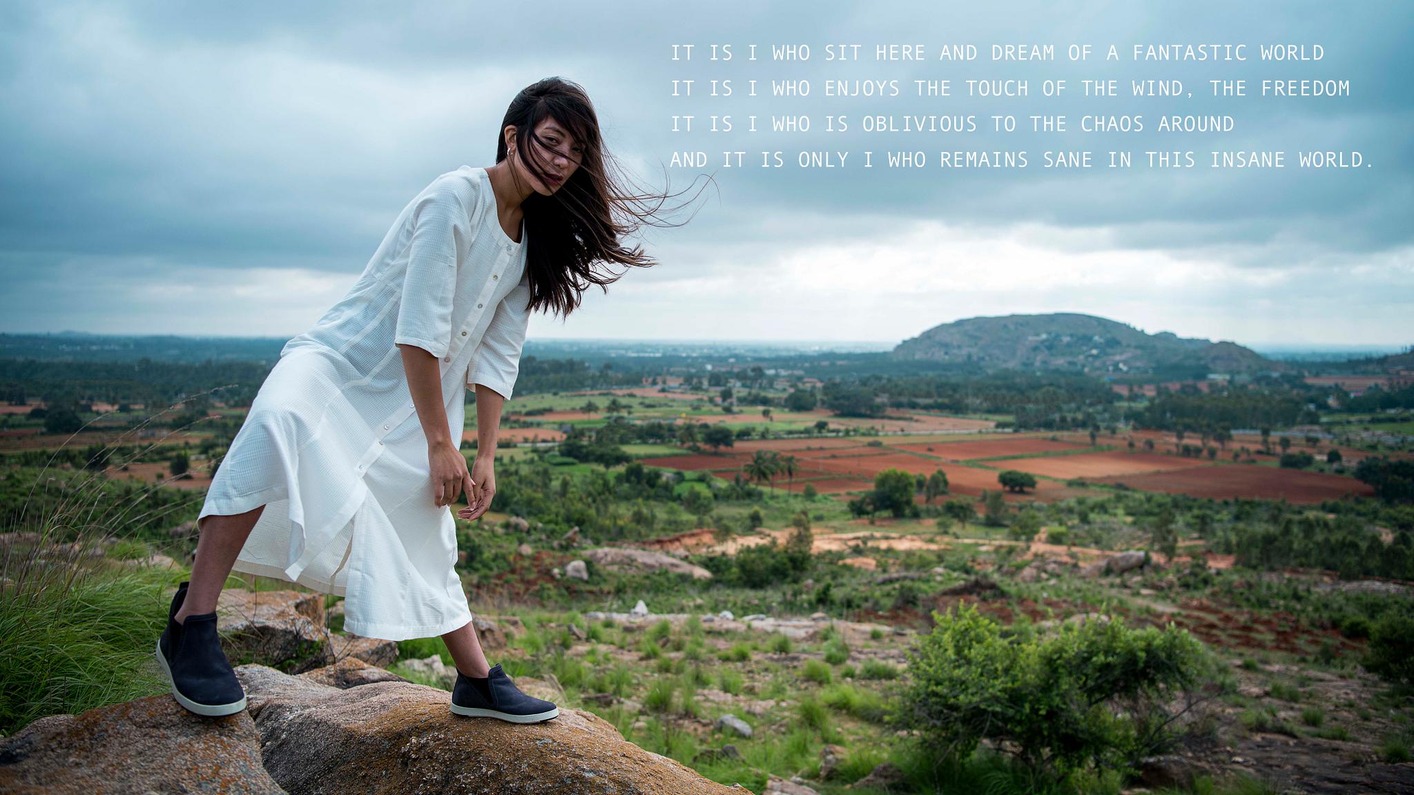 Person in white dress on a rocky hill with wind-blown hair, overlooking a vast rural landscape; overlaid with poetic text from 'The Parallel World' conceptual video series.