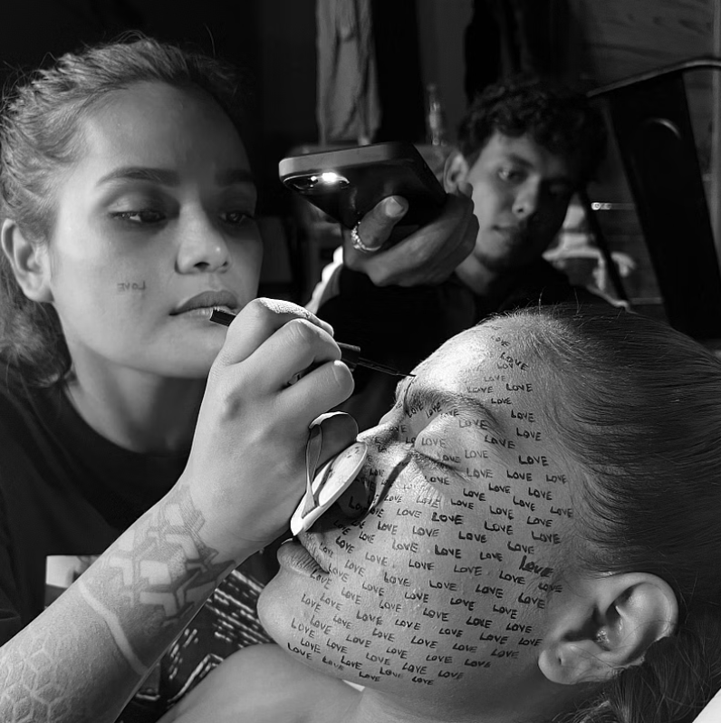Artist painting repeated “LOVE” text on a model’s face while another person lights the scene.