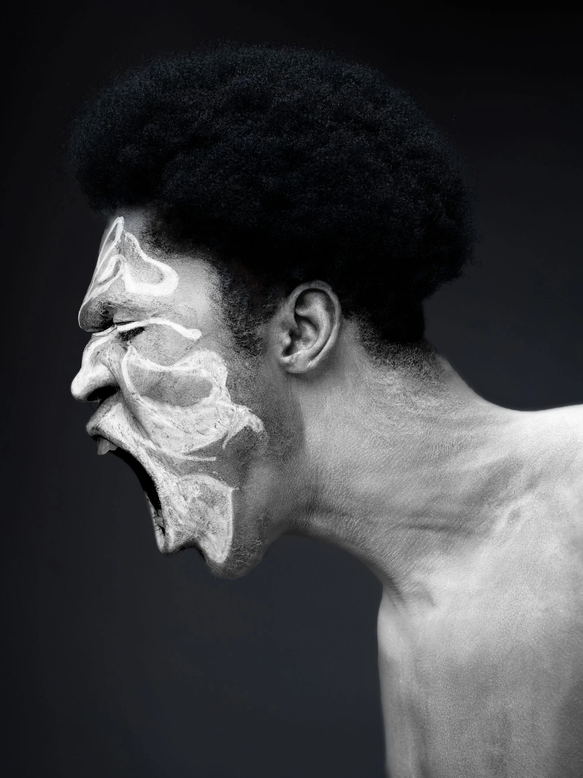 Side profile of a person with textured white face paint, captured mid-shout against a dark background.