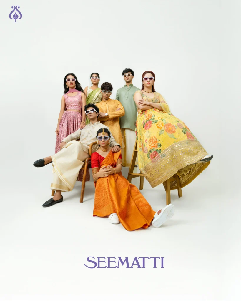 A group of seven young men and women dressed in traditional South Indian festive wear, styled with contemporary sunglasses. They strike confident, editorial-style poses in a studio setup with a white backdrop. The mix of silk sarees, lehengas, and kurta-pyjamas paired with modern accessories showcases a playful fusion of tradition and Gen Z attitude. The image is part of Seematti’s fashion campaign.