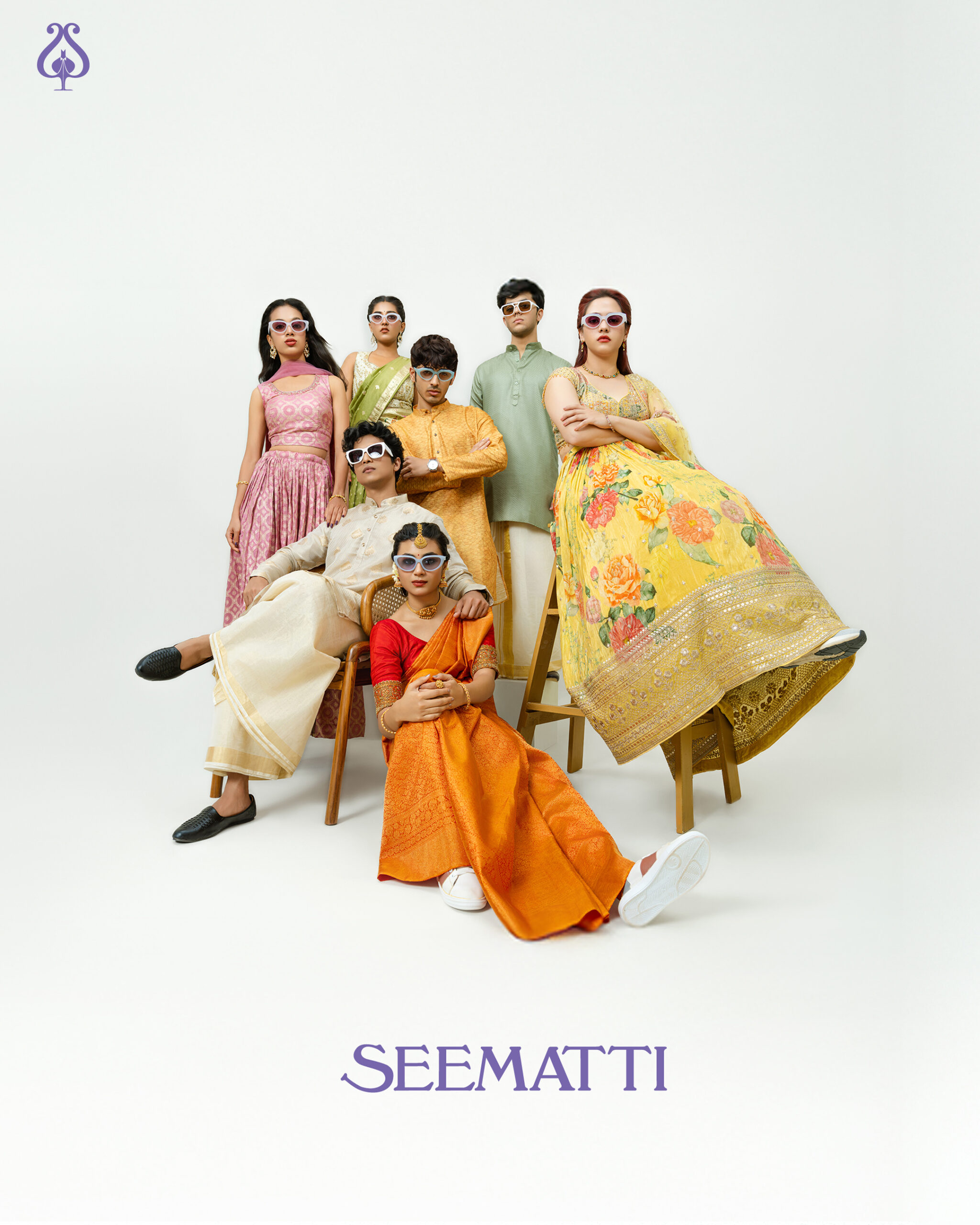 A group of seven young men and women dressed in traditional South Indian festive wear, styled with contemporary sunglasses. They strike confident, editorial-style poses in a studio setup with a white backdrop. The mix of silk sarees, lehengas, and kurta-pyjamas paired with modern accessories showcases a playful fusion of tradition and Gen Z attitude. The image is part of Seematti’s fashion campaign.