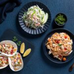 A spread of tacos, salad, and pasta arranged on a dark blue table.