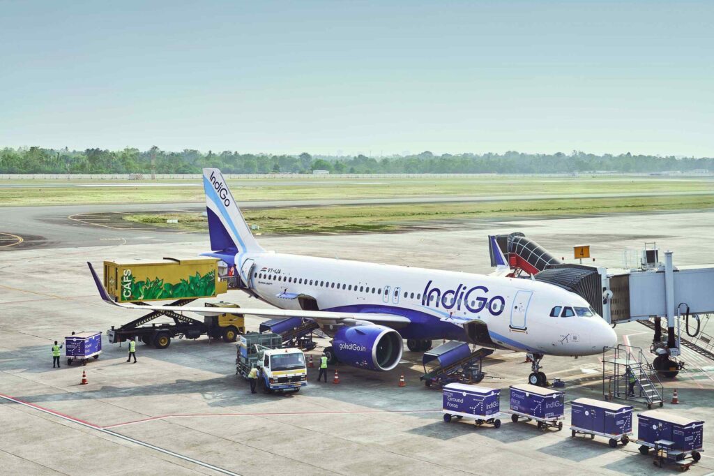 IndiGo aircraft parked at airport gate with ground crew loading luggage and cargo.