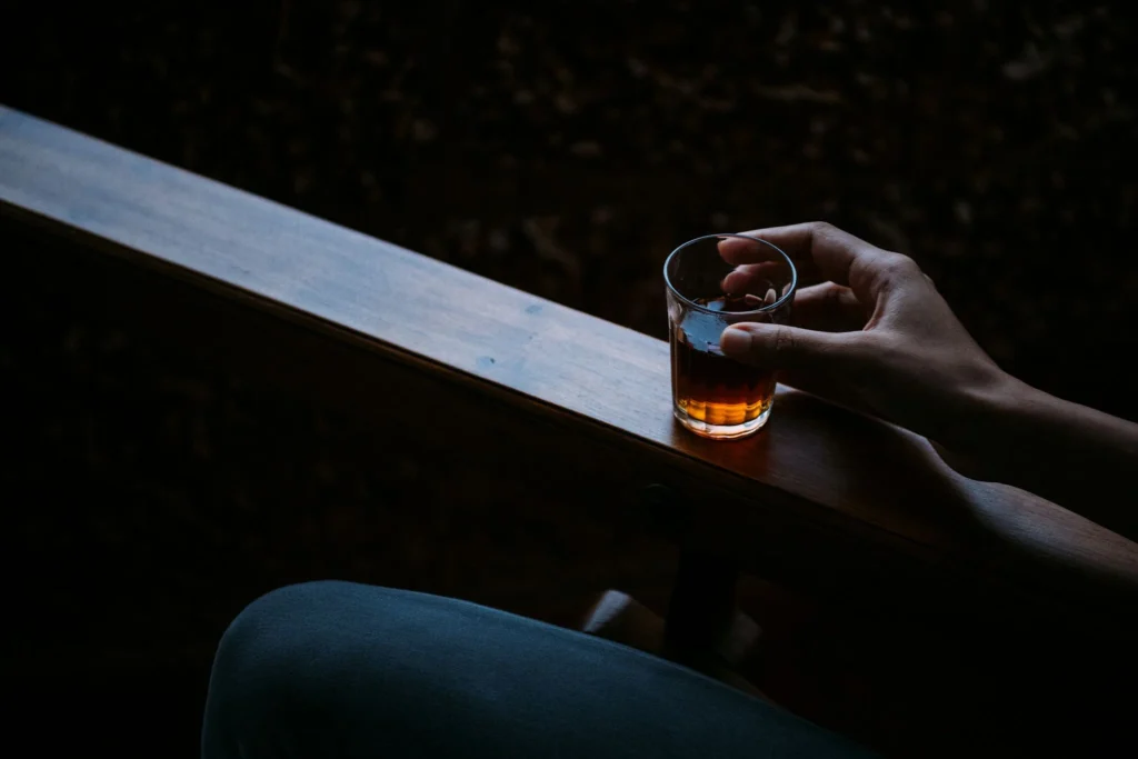 Close-up of a hand holding a glass of amber beverage on a wooden armrest in a dark, intimate setting.