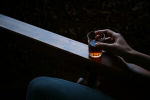 Close-up of a hand holding a glass of amber beverage on a wooden armrest in a dark, intimate setting.