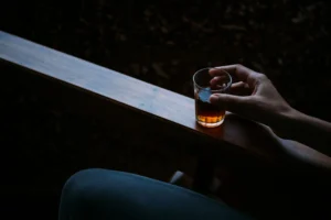 Close-up of a hand holding a glass of amber beverage on a wooden armrest in a dark, intimate setting.
