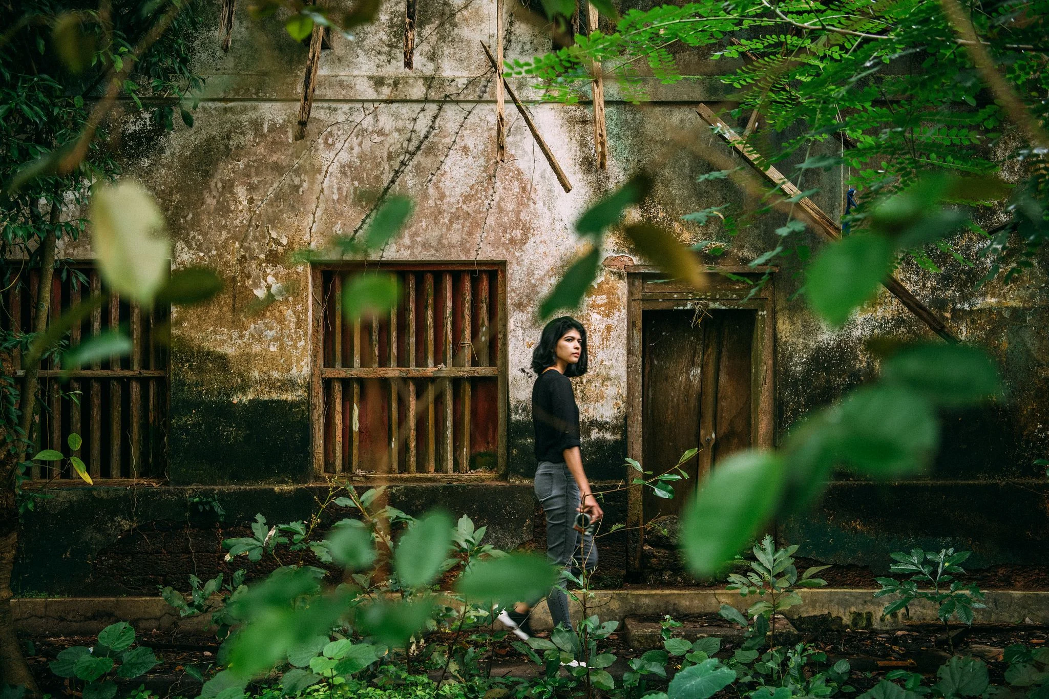 Woman walking past an abandoned, weathered building surrounded by dense green foliage and soft natural light.